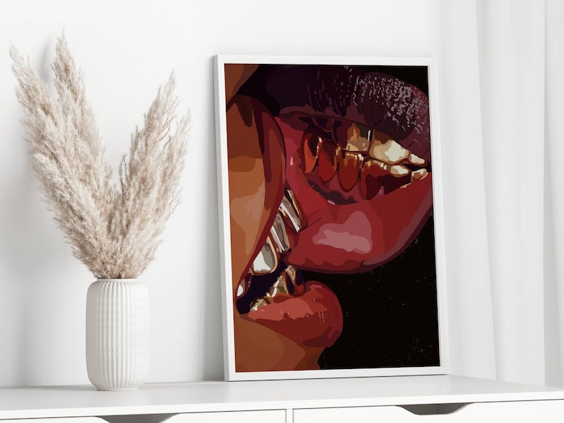 May include: A framed art print featuring a close-up of a mouth with red lips and gold teeth. The artwork has a dark background and is displayed on a white wall. A white vase with dried pampas grass is in the foreground.