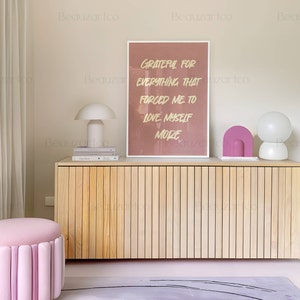 May include: A light wood cabinet with a pink and white lamp on top. The cabinet has a light wood finish and is made of vertical slats. The lamp is a modern design with a white shade and a pink base. The cabinet is sitting on a light gray rug with black lines. The wall behind the cabinet is a light beige color. A framed print with the text "Grateful for everything that forced me to love myself more" is hanging on the wall above the cabinet. A pink ottoman with a tufted design is in the foreground.