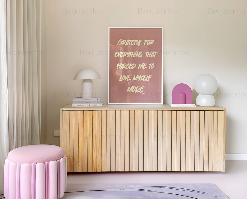 May include: A light wood cabinet with a pink and white lamp on top. The cabinet has a light wood finish and is made of vertical slats. The lamp is a modern design with a white shade and a pink base. The cabinet is sitting on a light gray rug with black lines. The wall behind the cabinet is a light beige color. A framed print with the text "Grateful for everything that forced me to love myself more" is hanging on the wall above the cabinet. A pink ottoman with a tufted design is in the foreground.