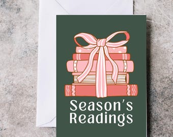 May include: A greeting card with a stack of pink books tied with a pink bow. The card has the words "Season's Readings" in white text on a dark green background. A white envelope is behind the card.