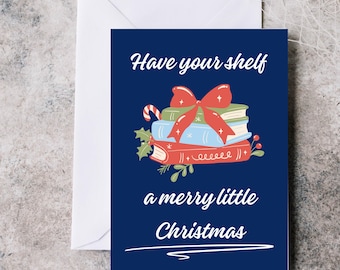 May include: A navy blue Christmas card with the text "Have your shelf a merry little Christmas." The card features a stack of books with a red bow, candy cane, and holly illustration. A white envelope is behind the card.