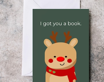 May include: A greeting card with a cartoon reindeer design. The reindeer has a red nose, blush, and a red scarf. The text "I got you a book" is at the top of the card. A white envelope is behind the card.