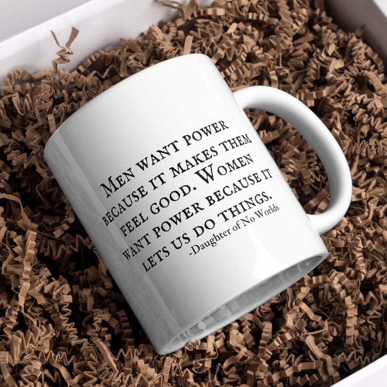 Puede incluir: Taza de cer&aacute;mica blanca con asa curva. La taza presenta un texto negro que dice: &laquo;LOS HOMBRES QUIEREN PODER PORQUE LES HACE SENTIR BIEN. LAS MUJERES QUIEREN PODER PORQUE NOS HACE HACER COSAS.&raquo; La cita se atribuye a &laquo;-Daughter of No Worlds&raquo;.