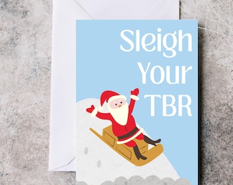 May include: A Christmas card with a cartoon Santa Claus sledding down a snowy hill. The card has a light blue background with the words "Sleigh Your TBR" in white. An envelope is behind the card.