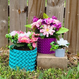 Puede incluir: Tres plantas en maceta con macetas de diferentes colores con patrones geométricos. La maceta más grande es turquesa, la maceta del medio es morada y la maceta más pequeña es gris. Las plantas están en flor con flores rosas, blancas y moradas.