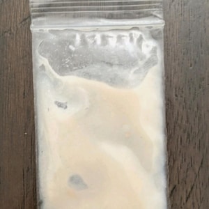 May include: A clear plastic bag filled with a light beige powder. The bag is sealed at the top with a zip closure. The bag is sitting on a dark brown wooden surface.