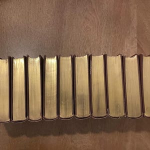 May include: A collection of hardcover books with gold-gilded edges. The spines are dark brown, and the pages have a golden hue. The books are arranged in a line on a wooden surface.