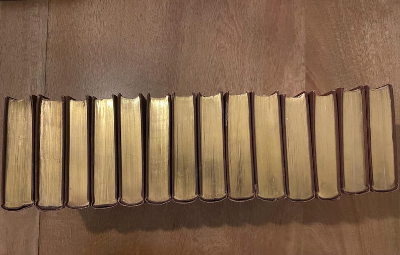 May include: A collection of hardcover books with gold-gilded edges. The spines are dark brown, and the pages have a golden hue. The books are arranged in a line on a wooden surface.
