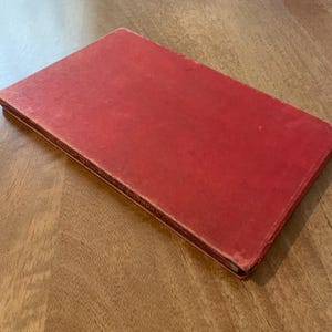 Op de afbeelding: Een gesloten, rood boek met een getextureerde kaft ligt op een houten oppervlak. De randen van het boek vertonen lichte slijtage, wat suggereert dat het vintage is. De titel van het boek is gedeeltelijk zichtbaar langs de rug.