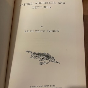 May include: An open book displaying the title "NATURE, ADDRESSES, AND LECTURES" by Ralph Waldo Emerson. The page is cream-colored with black text. A small illustration of a branch with leaves is printed on the page.