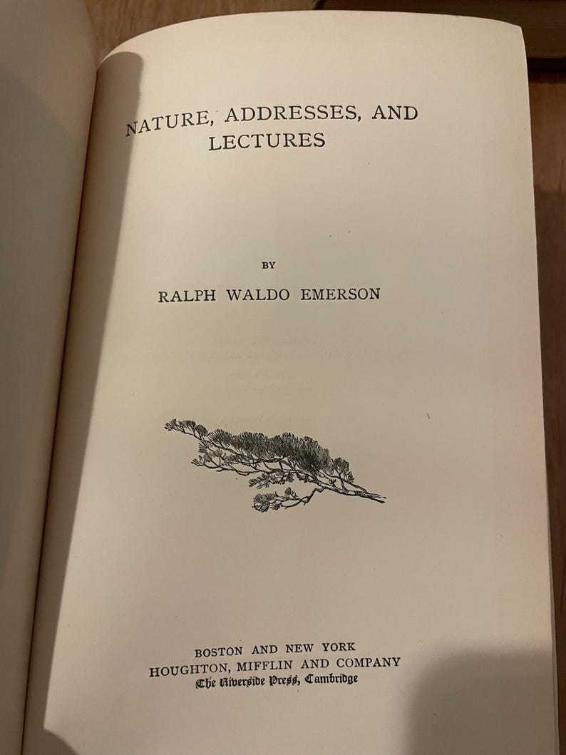 May include: An open book displaying the title "NATURE, ADDRESSES, AND LECTURES" by Ralph Waldo Emerson. The page is cream-colored with black text. A small illustration of a branch with leaves is printed on the page.