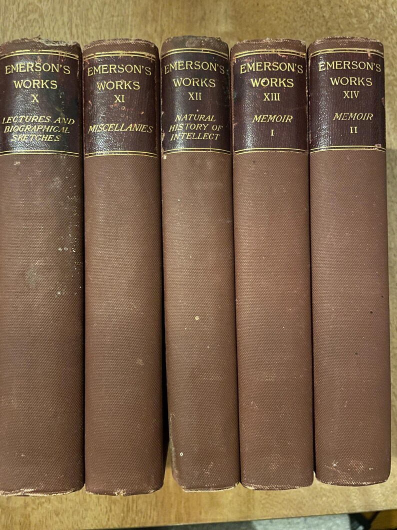 May include: A collection of five vintage books, each with a brown cloth cover and gold lettering. The spines read "Emerson's Works" with Roman numerals and titles such as "Miscellanies" and "Memoir". The books are arranged side by side.