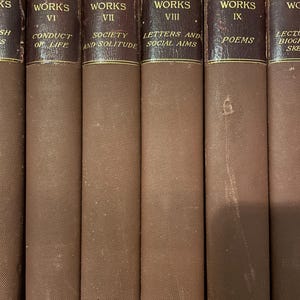 May include: A row of antique books, titled "Emerson's Works." The spines are a deep brown with gold lettering. Titles visible include "Conduct of Life," "Society and Solitude," "Letters and Social Aims," and "Poems."