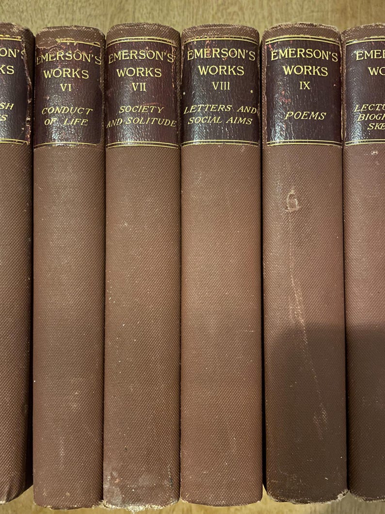 May include: A row of antique books, titled "Emerson's Works." The spines are a deep brown with gold lettering. Titles visible include "Conduct of Life," "Society and Solitude," "Letters and Social Aims," and "Poems."