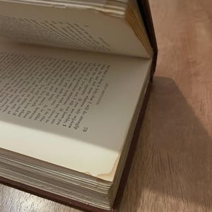 May include: An open antique book with aged, yellowed pages. The book has a dark brown cover and visible text on the pages. The corner of the page is slightly bent, showing signs of wear. The book is angled, revealing the text and the book's binding.
