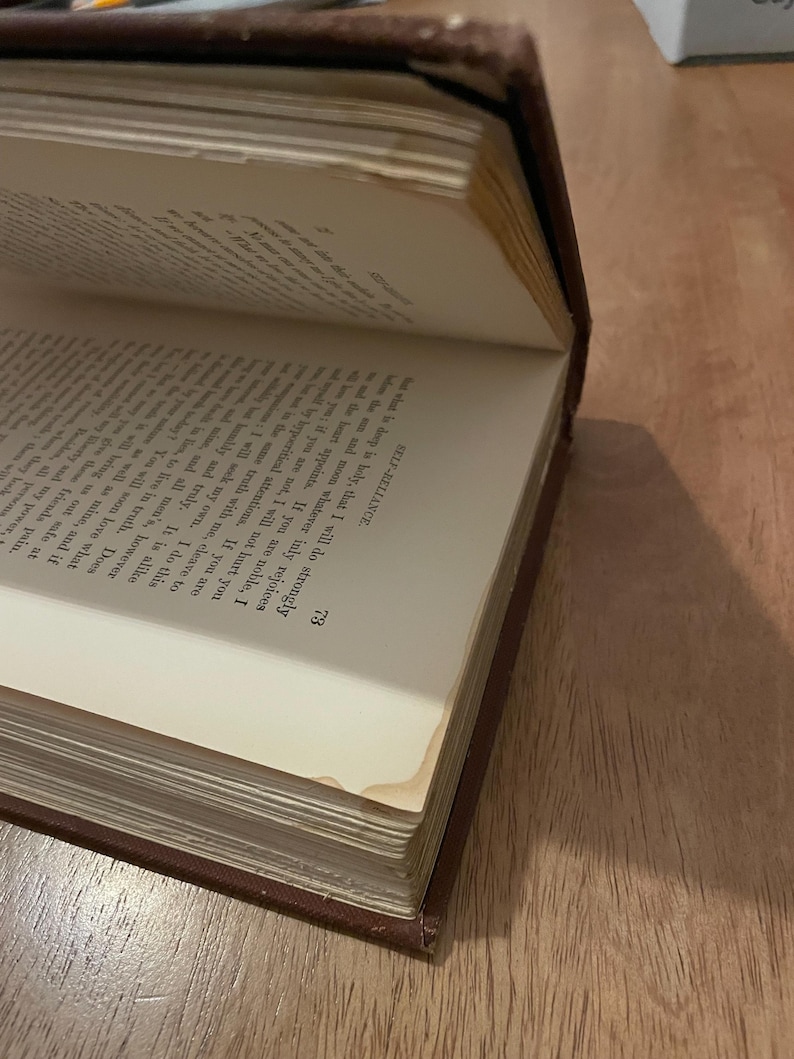 May include: An open antique book with aged, yellowed pages. The book has a dark brown cover and visible text on the pages. The corner of the page is slightly bent, showing signs of wear. The book is angled, revealing the text and the book's binding.