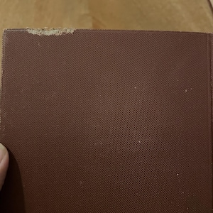 May include: Close-up of a burgundy book cover with a textured pattern. The top corner of the book shows some wear and tear. The book's cover is a solid, dark red-brown colour.