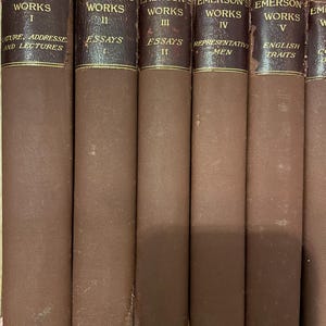 May include: A row of vintage books with brown fabric covers. The spines are dark brown with gold lettering, displaying "EMERSON'S WORKS" and volume numbers. Titles include "Nature, Addresses and Lectures," "Essays," "Representative Men," and "English Traits."