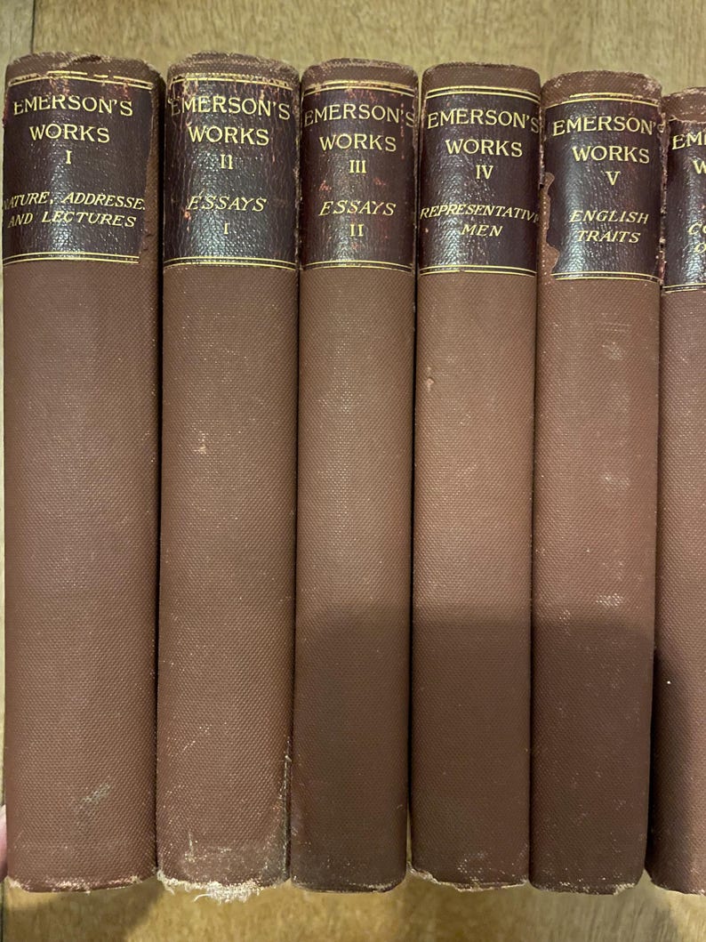 May include: A row of vintage books with brown fabric covers. The spines are dark brown with gold lettering, displaying "EMERSON'S WORKS" and volume numbers. Titles include "Nature, Addresses and Lectures," "Essays," "Representative Men," and "English Traits."