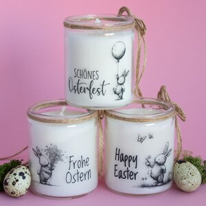 Könnte beinhalten: Drei weiße Kerzen in Gläsern mit Kordeldetails, jede mit einem Ostermotiv. Eine Kerze zeigt ein Häschen mit einem Ballon, eine andere ein Häschen mit Blumen und die letzte ein Häschen mit einer Blume und Schmetterlingen. Der Text auf den Kerzen lautet "Schönes Osterfest", "Frohe Ostern" und "Happy Easter".