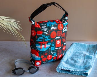 Peut inclure: Sac à motifs noirs et rouges avec une poignée et une boucle noires, orné d'illustrations sur le thème du Japon. Une serviette bleu clair et des lunettes de natation noires sont à côté du sac.