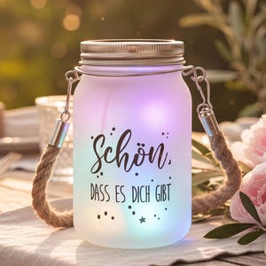 Può includere: Un barattolo di vetro smerigliato con coperchio in metallo e manico in corda, illuminato con luci colorate. Il barattolo ha il testo "Schön, dass es dich gibt" in scrittura nera. Il barattolo è su un tavolo con fiori.