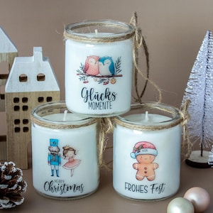 Può includere: Tre candele bianche in barattoli di vetro con illustrazioni e testo festivi. Una candela ha gufi, un'altra uno schiaccianoci e una ballerina, e l'ultima un omino di pan di zenzero. I barattoli sono decorati con spago.