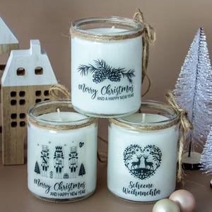 Può includere: Tre candele bianche in barattoli di vetro con disegni natalizi. Le candele presentano pigne, schiaccianoci e cervi. Ogni barattolo è avvolto con spago e ha una candela bianca all'interno. Alberi e case natalizie decorative sullo sfondo.