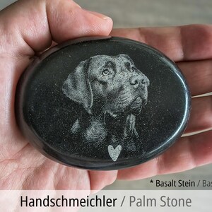 Pu&ograve; includere: Una pietra da palmo in basalto nero, liscia e di forma ovale, con il ritratto di un cane inciso al laser e un piccolo simbolo a forma di cuore sotto il mento. La pietra presenta una superficie lucida e riflettente di colore dal grigio scuro al nero.