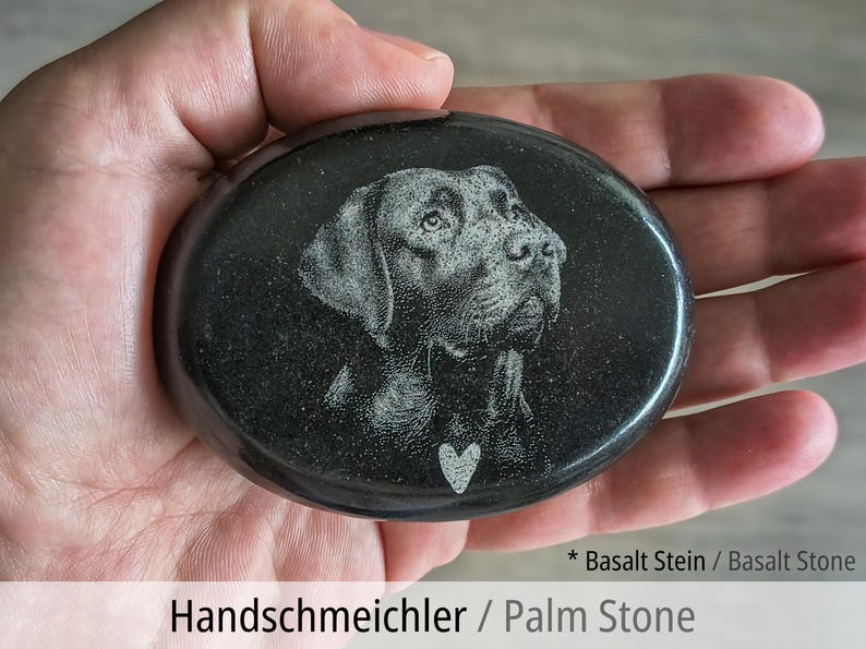 Pu&ograve; includere: Una pietra da palmo in basalto nero, liscia e di forma ovale, con il ritratto di un cane inciso al laser e un piccolo simbolo a forma di cuore sotto il mento. La pietra presenta una superficie lucida e riflettente di colore dal grigio scuro al nero.