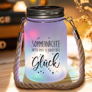 Può includere: Un barattolo di vetro smerigliato con coperchio in metallo e manico in corda, illuminato da luci colorate. Il barattolo presenta il testo tedesco "Sommernächte...Glück". Un oggetto decorativo.