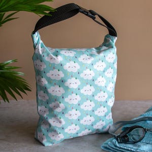 Puede incluir: Una bolsa impermeable azul claro con asa negra y cierre de hebilla. La bolsa tiene un estampado de nubes blancas sonrientes y gotas de lluvia. Una toalla azul y gafas de natación están junto a la bolsa.
