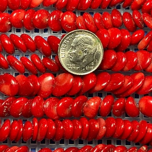 以下が含まれることがあります： 鮮やかな赤色のサンゴビーズのストランドのクローズアップ。ビーズは楕円形で、つながっています。サイズの比較のために、米国の10セント硬貨がビーズの上に置かれています。ビーズは白いグリッド上にあります。