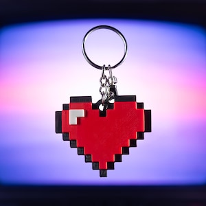 May include: A pixelated red heart keychain with a silver keyring and chain. The heart design features black and white accents, reminiscent of retro video game graphics. The keychain is suspended against a blurred, colorful background.