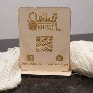 May include: A wooden sign with the text "Stellar Knits" and a yarn ball logo. The sign also includes a QR code, a Facebook icon with the text "SandSknits", and an Instagram icon with the text "Stellarknitsco".