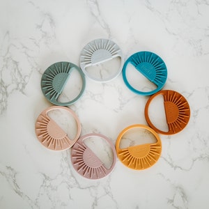 May include: Eight silicone teething toys in a circle on a white marble surface. The toys are shaped like suns with a half-circle cut out. The colors are light blue, teal, light pink, peach, orange, brown, and yellow.