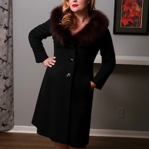 May include: A black wool coat with a flared skirt and a luxurious brown fur collar. The coat features gold buttons and pockets. A black veil is worn on the head. The coat is a classic vintage style.