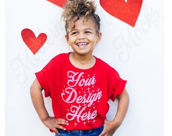 Puede incluir: Un niño pequeño con una camiseta roja con el texto "Your Design Here" en rosa. El niño sonríe y está de pie frente a una pared blanca con corazones de papel rojo.