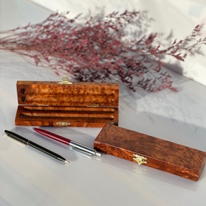 May include: Two wooden boxes with brass clasps, one open and one closed, with two pens, one black and one red, on a white surface.