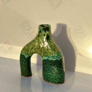 Può includere: Un vaso in ceramica verde e oro con un'apertura a forma di arco. Il vaso ha una smaltatura macchiettata e un design astratto unico.
