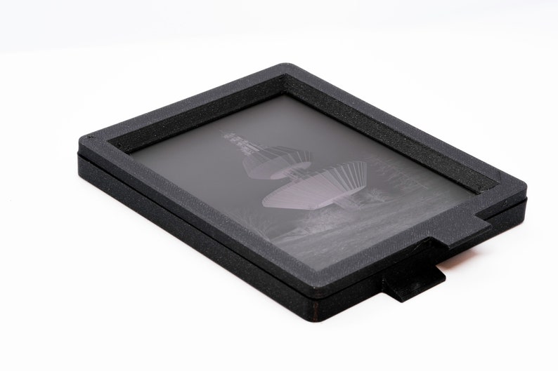 May include: A black frame with a glass insert, holding a vintage photographic negative of a structure with a circular design.