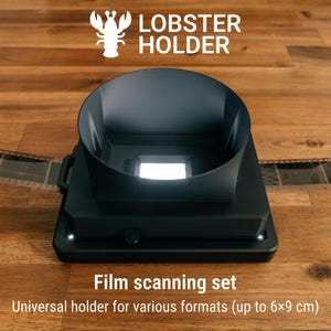 May include: Black film scanning set with the text "LOBSTER HOLDER". The set includes a universal holder for various film formats up to 6x9 cm. The set is on a wooden surface.