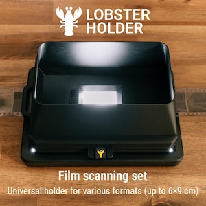 Może przedstawiać: Czarny zestaw do skanowania filmów Lobster Holder z prostokątną podstawą i centralnym otworem świetlnym. Urządzenie posiada małą żółtą ikonę homara na przednim zatrzasku i trzyma pasek filmu 35mm. Tekst: Lobster Holder oraz Film scanning set.