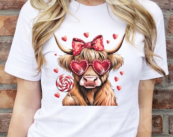 Pode incluir: Camiseta branca com uma ilustração de desenho animado de uma vaca Highland usando óculos de sol em forma de coração vermelho e um laço vermelho e branco de bolinhas. A vaca está segurando uma pirulito vermelho e branco.