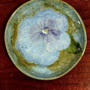 Op de afbeelding: Een rond keramieken bord met een handgeschilderde lichtblauwe bloem met donkerblauwe nerven in het midden. De achtergrond van het bord vertoont een gevlekte mix van aardse groen-, bruin- en blauwtinten met een glanzende en gespikkelde afwerking.