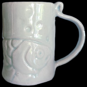 May include: A handmade ceramic mug in a light blue glaze. The mug features an embossed fish design and bubble-like textures. The handle has a unique, curved shape. The mug is suitable for hot or cold beverages.
