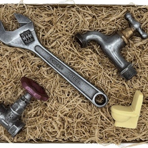 Op de afbeelding: Een cadeaudoos gevuld met bruin verpakkingsmateriaal en vier chocolade replica's van loodgietersgereedschap: een moersleutel, een kraan, een pijpventiel en een toilet. De moersleutel is gemarkeerd met "FORGED STEEL".
