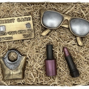 May include: A gift box filled with brown shredded paper and containing a gold credit card with the text "CREDIT CARD" and "I'M RICH", a pair of gold sunglasses, a gold handbag, a dark pink nail polish bottle, and a black lipstick.