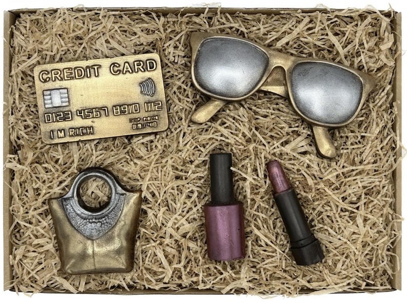 May include: A gift box filled with brown shredded paper and containing a gold credit card with the text "CREDIT CARD" and "I'M RICH", a pair of gold sunglasses, a gold handbag, a dark pink nail polish bottle, and a black lipstick.