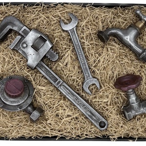 Op de afbeelding: Een set van vijf metalen gereedschappen in een doos gevuld met bruin papier. De gereedschappen omvatten een sleutel, een pijpsleutel, een kraan, een klep en een sleutel met de woorden "DROP FORGED" en "SUPER LEGGO" erop.